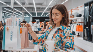 Texto alternativo: Mujer sonriente con gafas y camisa colorida sosteniendo bolsas de compra en una tienda, representando la experiencia del cliente en marketing y publicidad.