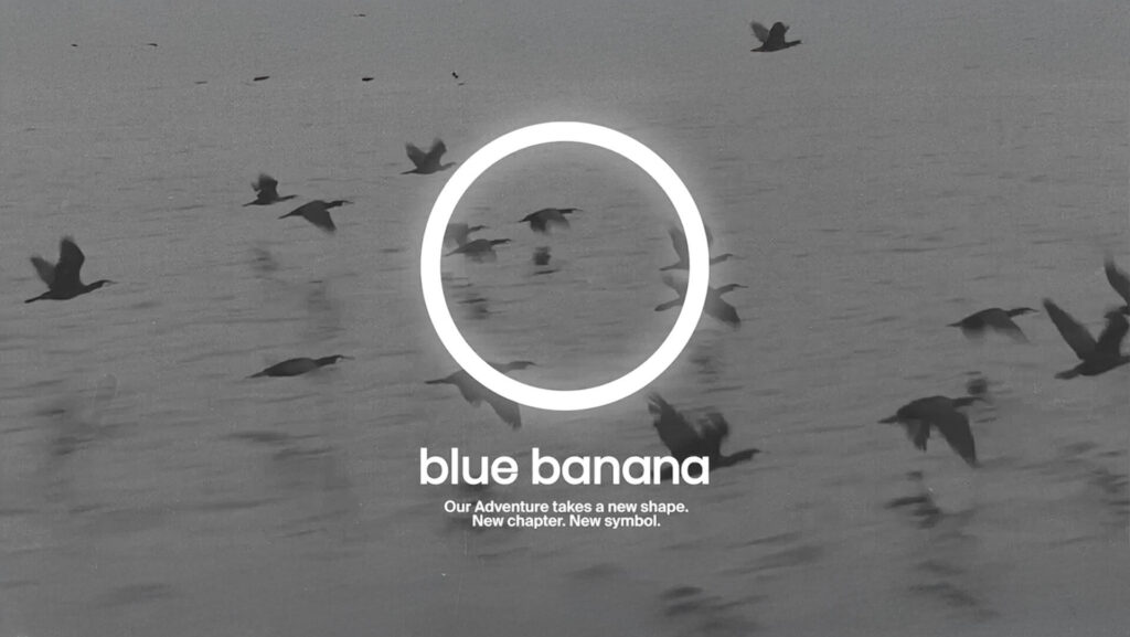 Logotipo circular blanco de Blue Banana sobre una imagen en blanco y negro de aves volando sobre el agua, acompañado del mensaje “Our Adventure takes a new shape. New chapter. New symbol.” que refuerza su proceso de rebranding.