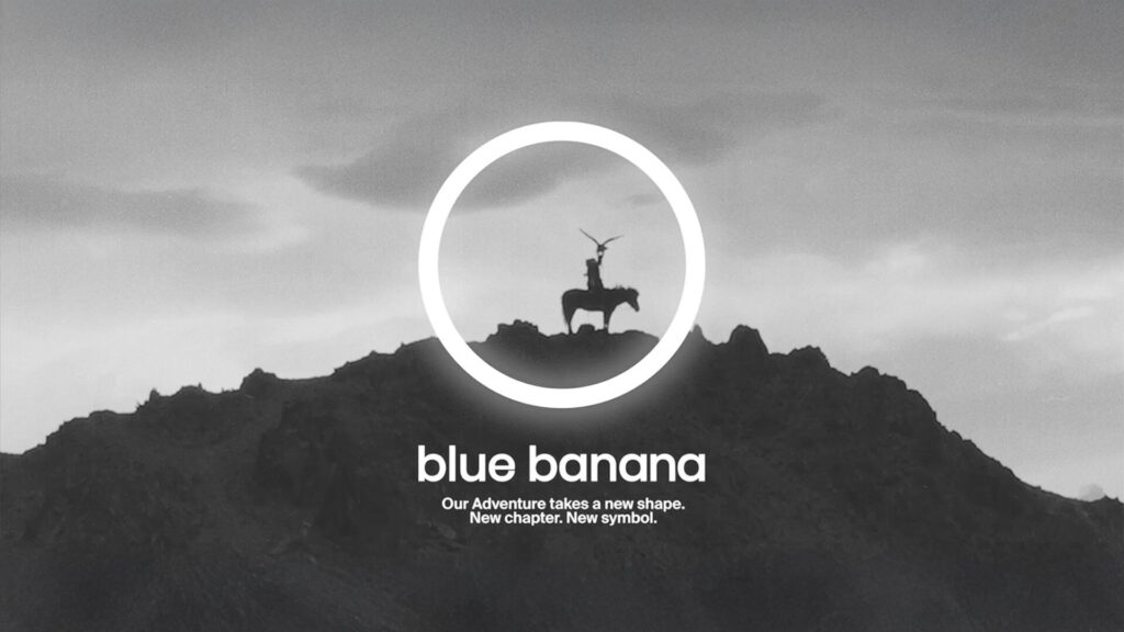 Nuevo logotipo circular de Blue Banana en blanco sobre fondo gris con silueta de jinete en la cima de una montaña, acompañado del mensaje “Our Adventure takes a new shape. New chapter. New symbol.” que representa el rebranding de la marca.