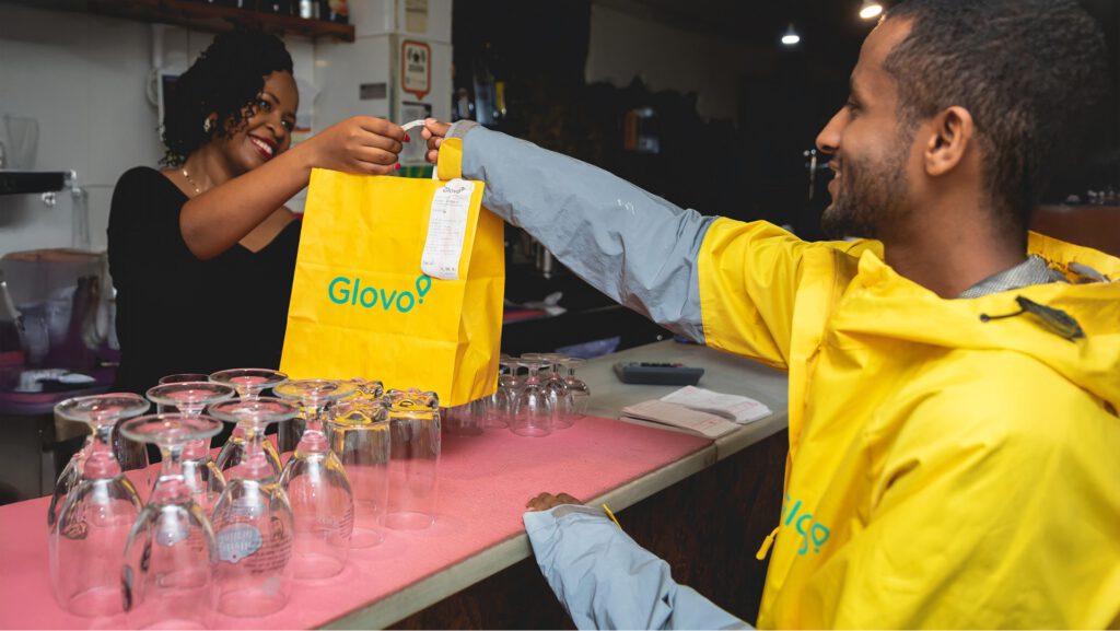 Repartidor de Glovo Delivery recogiendo un pedido en un restaurante mientras una trabajadora le entrega la bolsa amarilla.