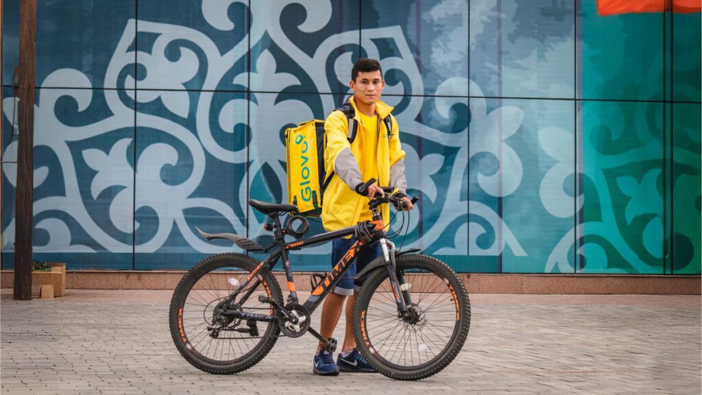 Repartidor de Glovo Delivery con mochila amarilla posando junto a su bicicleta frente a una pared decorada.
