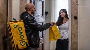 Repartidor de Glovo Delivery entregando una bolsa amarilla a una clienta en la puerta de su casa.