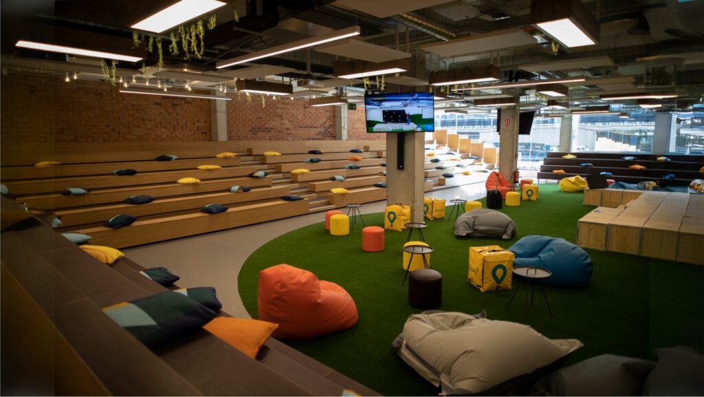 Espacio de trabajo colaborativo en las oficinas de Glovo Delivery con sillas, mesas altas y decoración en tonos verdes y amarillos.